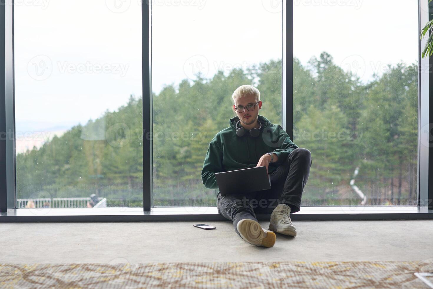 A blonde man sitting on the floor of a modern office, leaning against the window with a laptop in his lap, fully engaged in his work while maintaining a relaxed and focused demeanor. photo