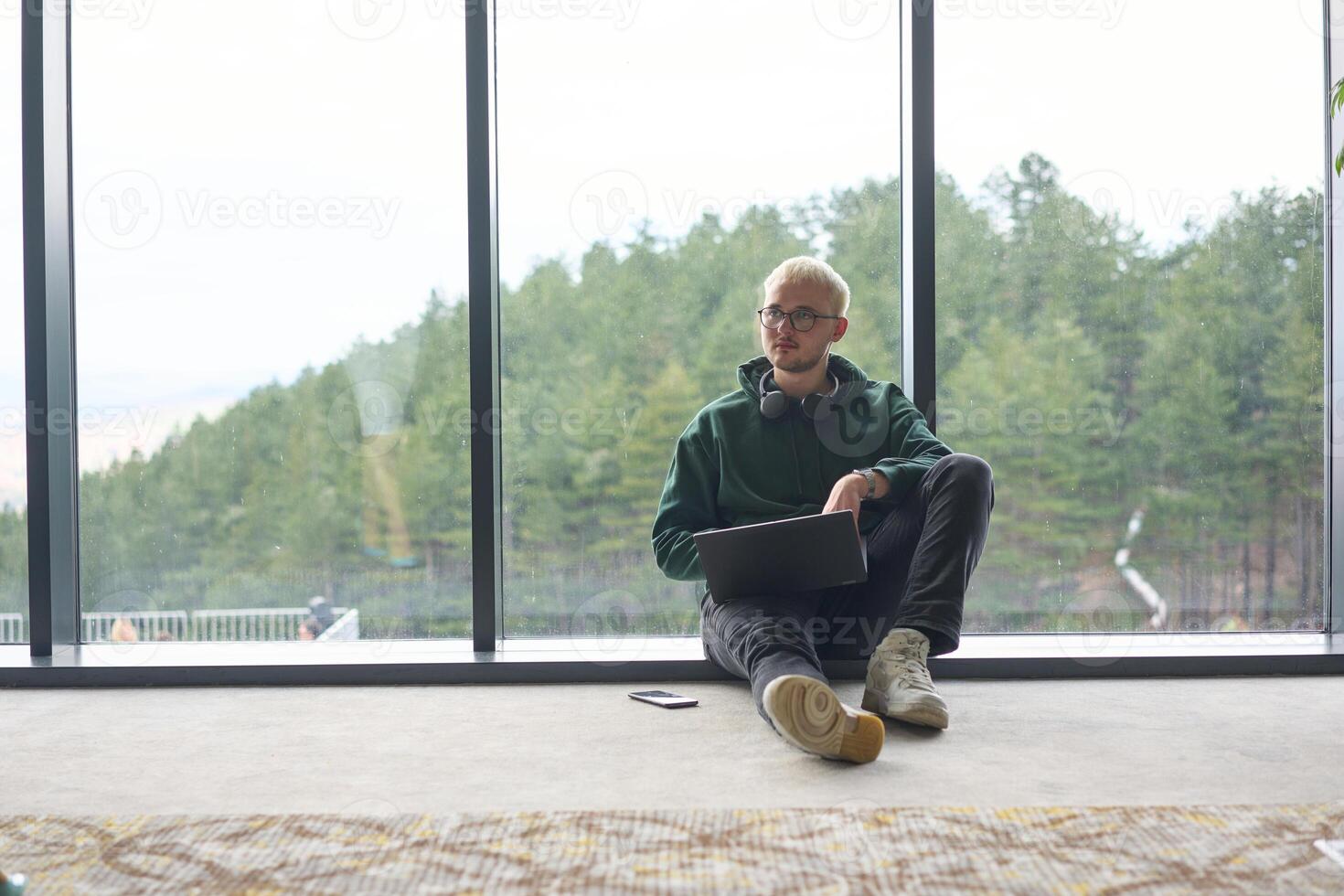 A blonde man sitting on the floor of a modern office, leaning against the window with a laptop in his lap, fully engaged in his work while maintaining a relaxed and focused demeanor. photo