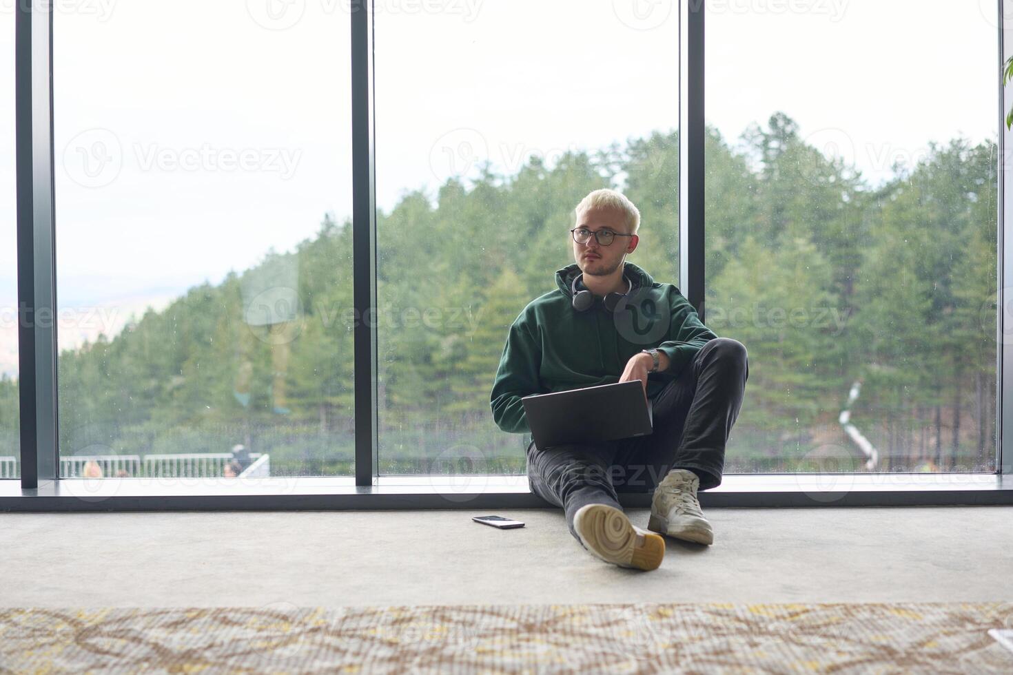 A blonde man sitting on the floor of a modern office, leaning against the window with a laptop in his lap, fully engaged in his work while maintaining a relaxed and focused demeanor. photo