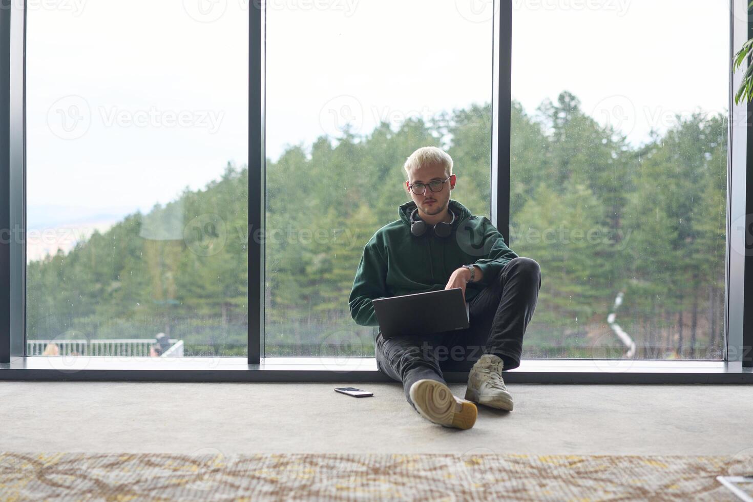 A blonde man sitting on the floor of a modern office, leaning against the window with a laptop in his lap, fully engaged in his work while maintaining a relaxed and focused demeanor. photo