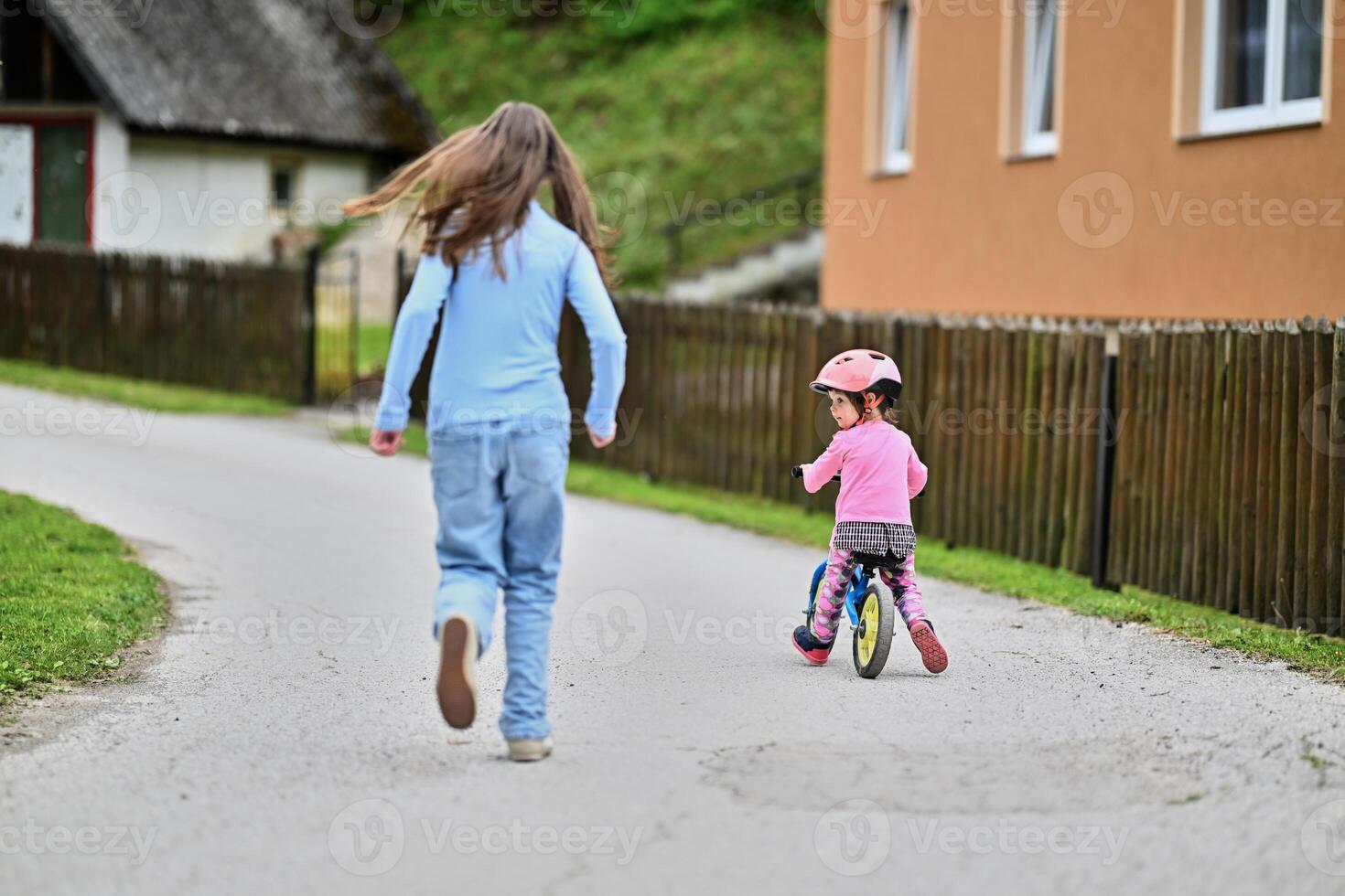 A joyful teenager encourages her younger sister as she learns to ride a bike, both sharing a moment of laughter and bonding in the sunny outdoors photo