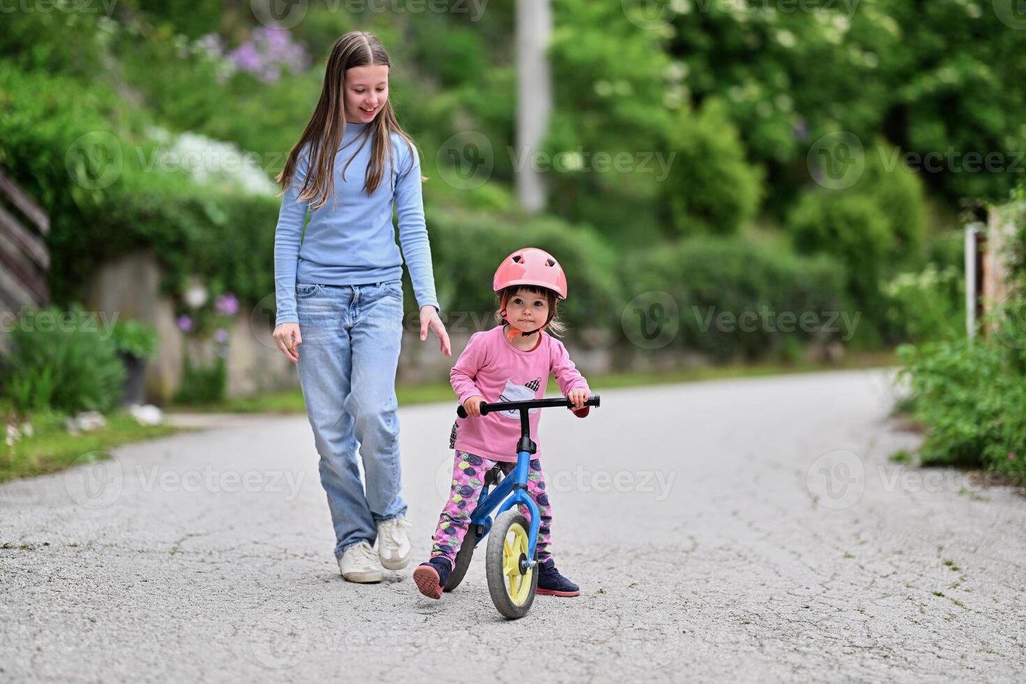 A joyful teenager encourages her younger sister as she learns to ride a bike, both sharing a moment of laughter and bonding in the sunny outdoors photo