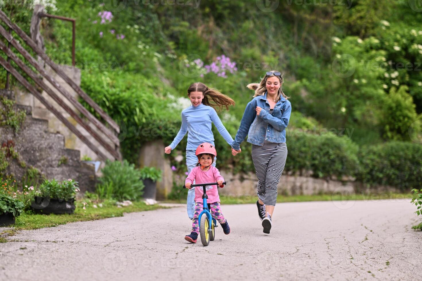 A cheerful little girl riding a balance bike with a helmet while two women joyfully run beside her on a paved path in a lush outdoor setting. photo