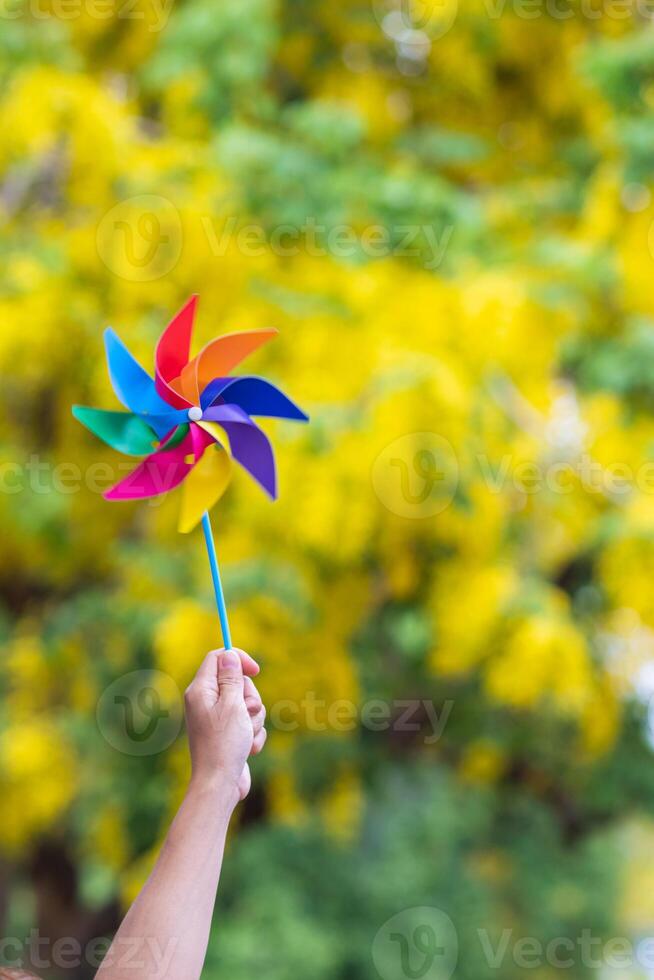 girl's hands raised above her head holding rainbow-colored windmill looked beautiful against bright yellow flower background. toy windmill in girl's hand on yellow blur background looks beautiful. photo