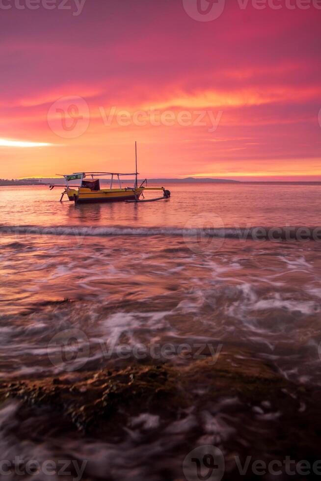 A fisherman's boat rests on the shore at Pasir Panjang Beach during sunset. The sky is adorned with clouds in beautiful red and yellow gradients, creating a stunning and tranquil scene photo