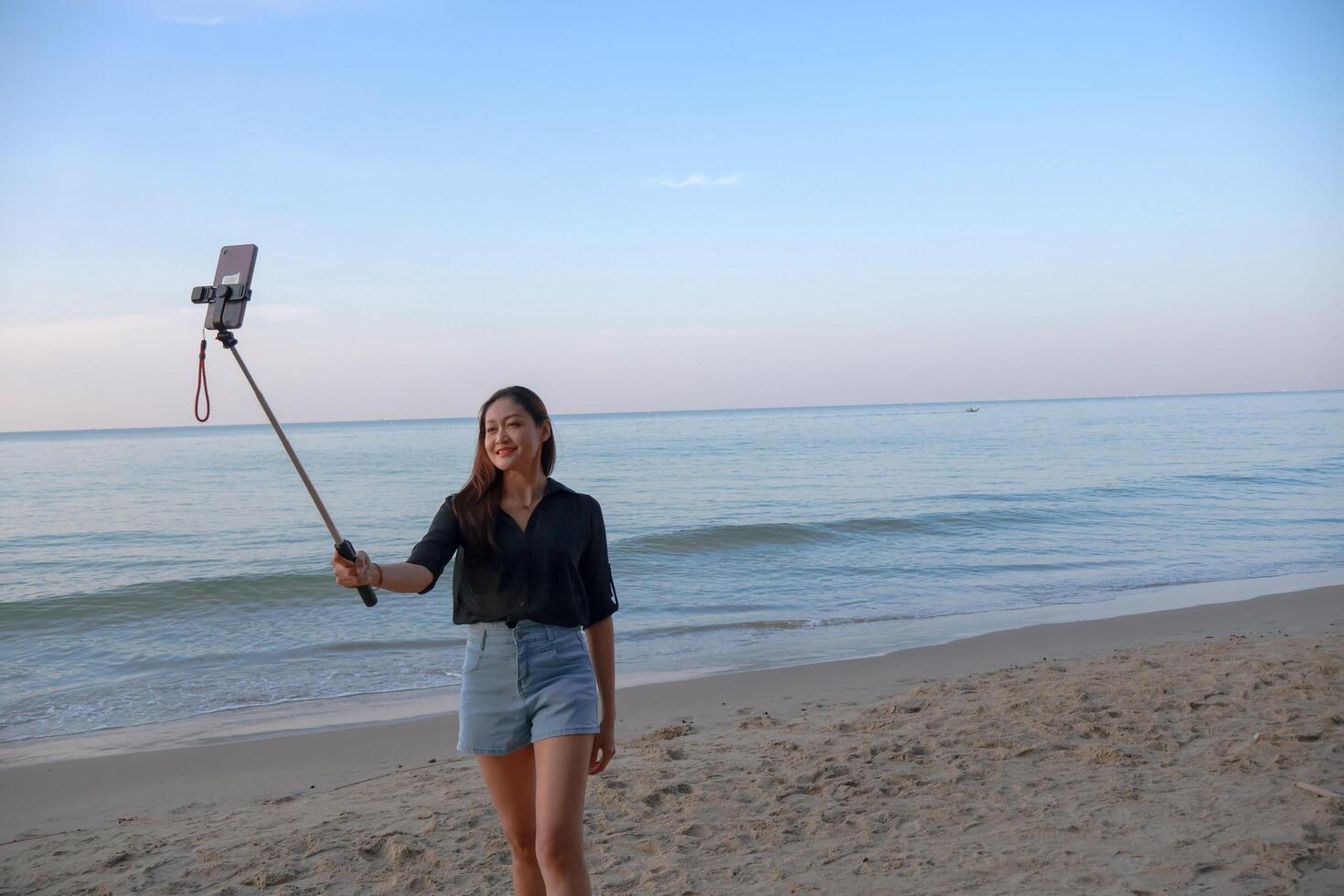 Asian woman a selfie stick and using a smartphone to take photographs by the sea. photo