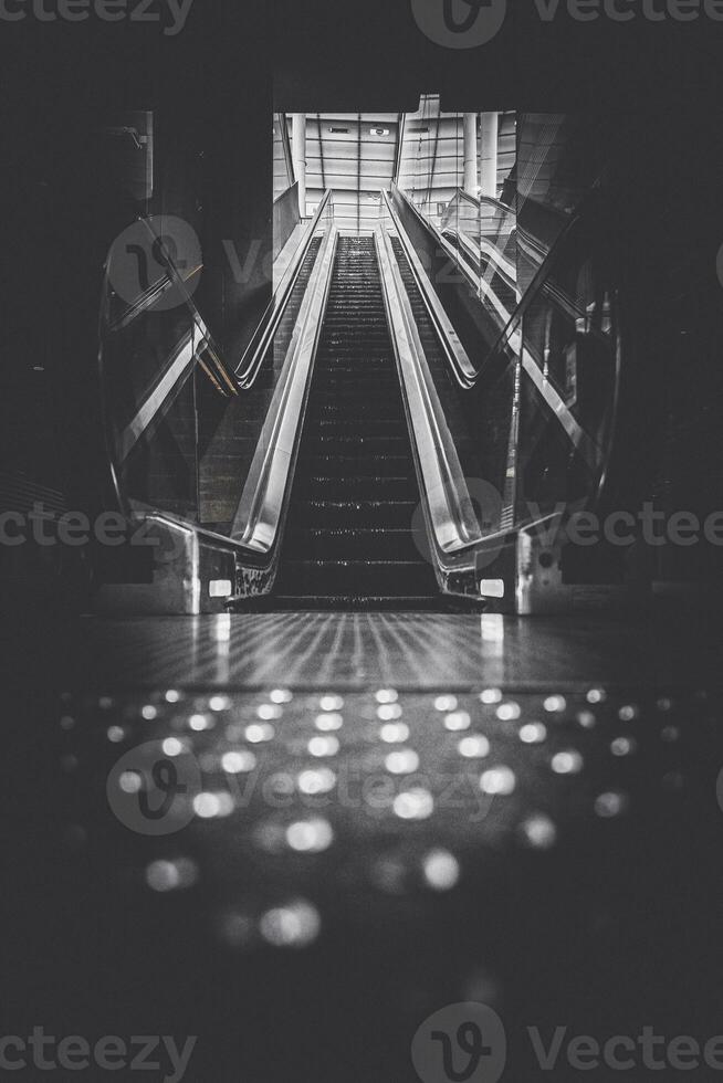Black and white vertical shot a dimly lit escalator leading to a bright ceiling photo