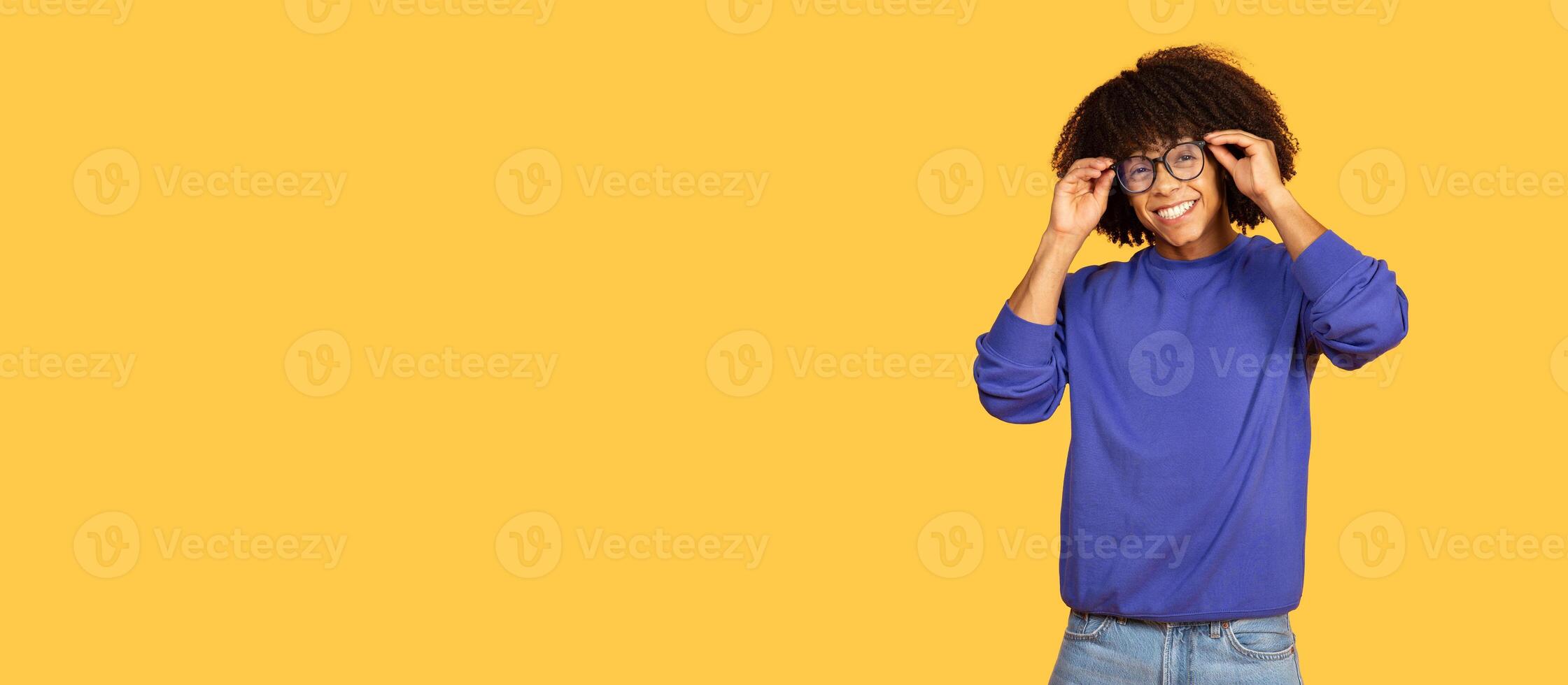A joyful young black man with curly hair adjusts his glasses while smiling. He wears a casual blue sweater and stands in front of a bright yellow wall, copy space photo