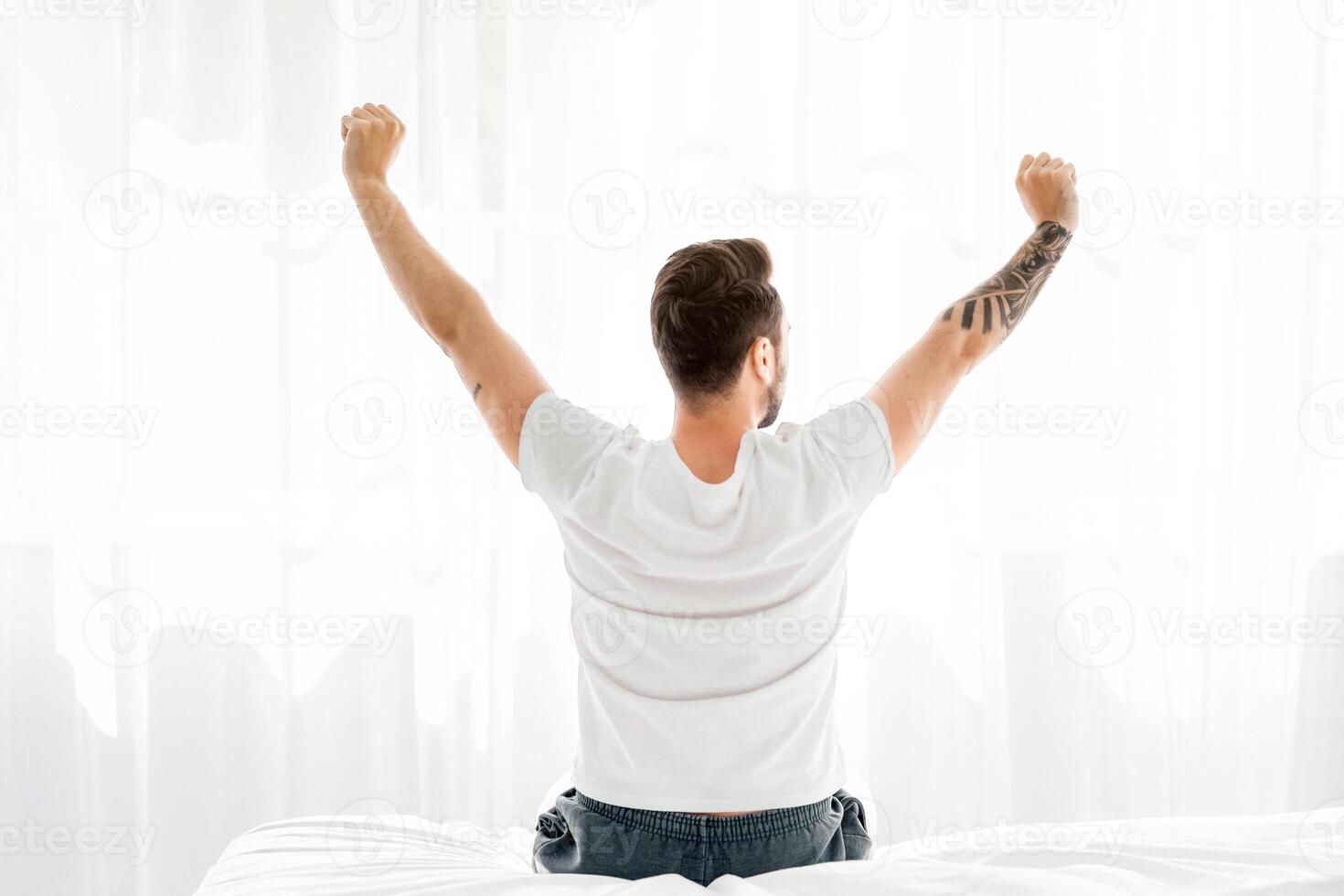 Relaxed young man waking up in bed and stretching his arms, facing the window, back view, free space. Millennial guy enjoying morning and new day photo