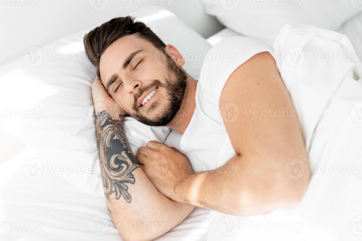 Happy morning. Satisfied guy waking up, feeling rested, happy bearded black guy lying on white linen flax bedsheets and smiling, relaxing in bedroom photo