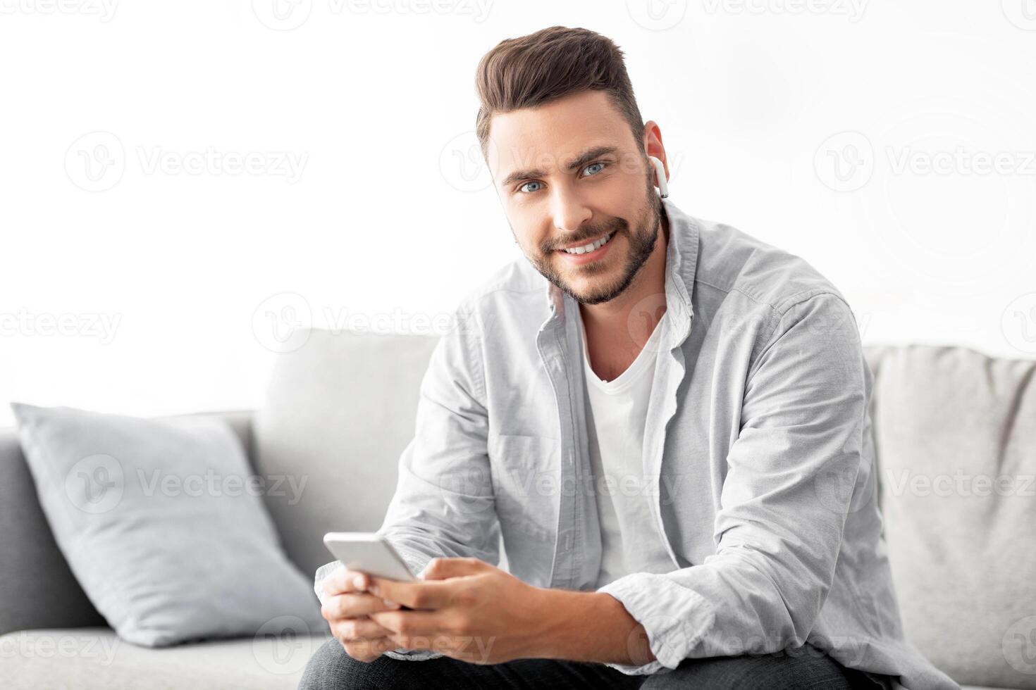 Handsome millennial man in earbuds using smartphone and smiling to camera, sitting on sofa at home, copy space. Happy guy texting on cellphone in living room interior photo