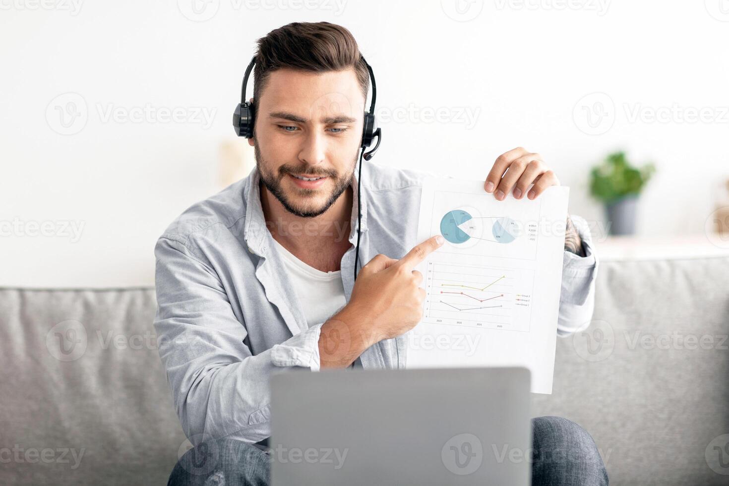 Online meeting. Happy young manager having call on laptop computer, showing diagram and market reports to webcam. Man wearing headset and looking at pc screen photo