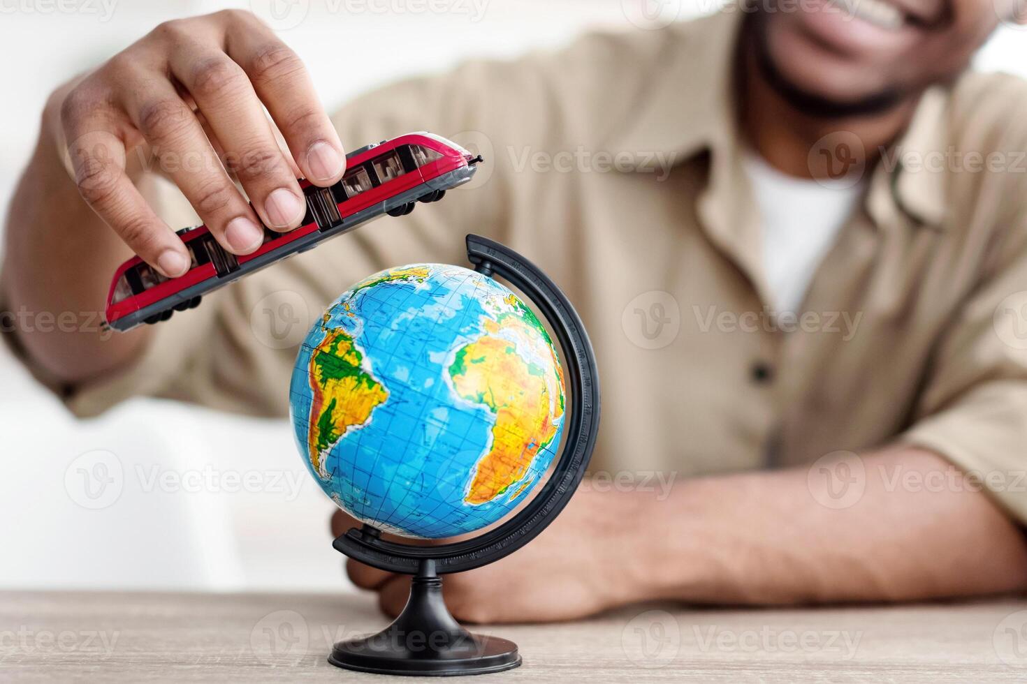 Travel Concept. Black Man Playing With Train Model And World Globe Sitting At Desk At Home, Planning Summer Vacation, Dreaming About Trip. Railway Tickets Concept. Selective Focus On Toy photo