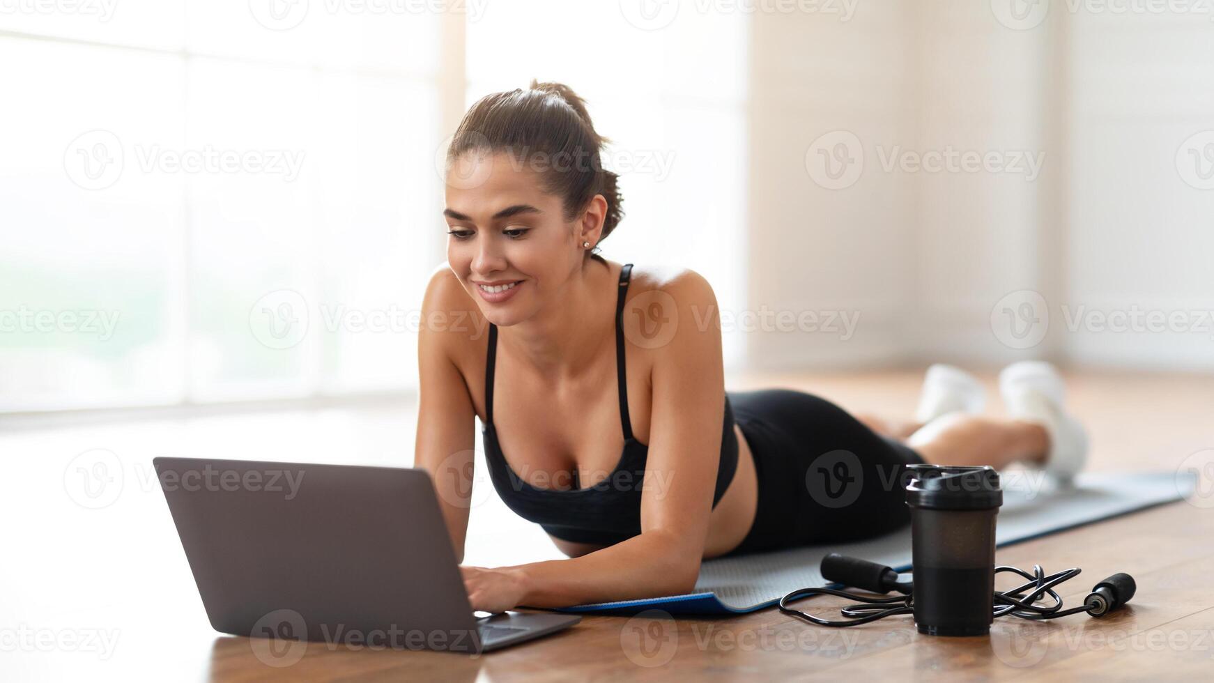 Sporty young lady exercising at home, using pc choosing lesson on laptop, watching fitness on Internet, happy female having online virtual class in living room, typing on keyboard photo