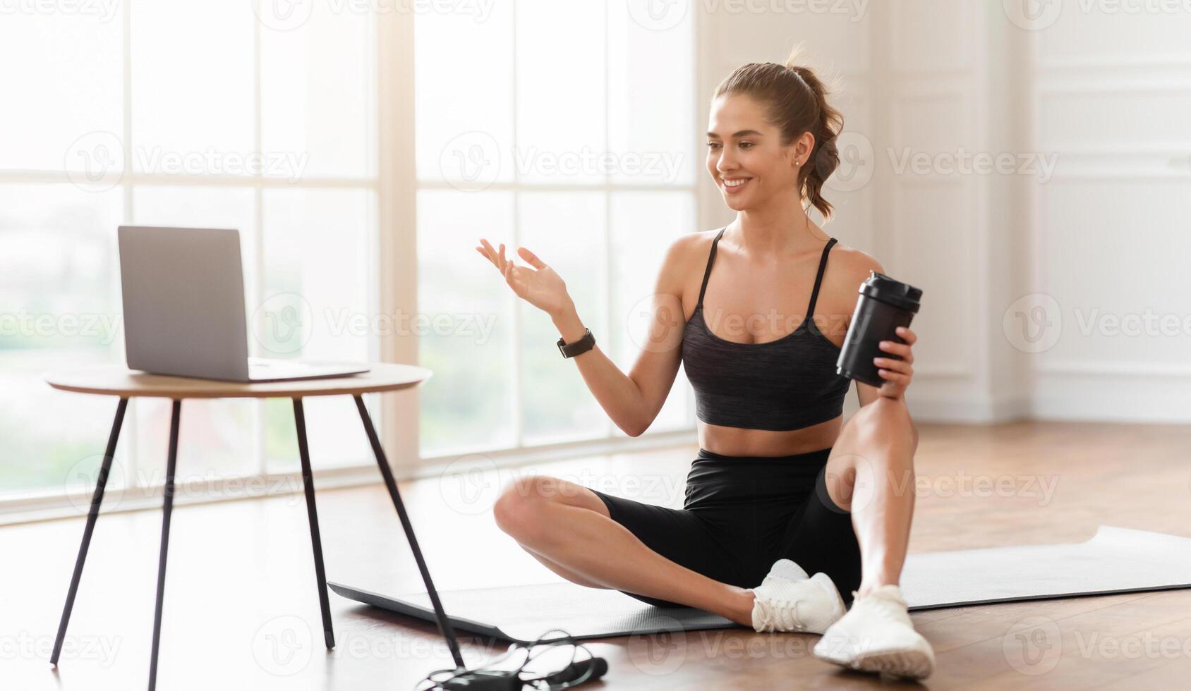 Online Exercise Session Marathon, Remote Training. Happy millennial female sitting on floor mat and talking with coach, having videocall with teacher, using pc for virtual workout, drinking water photo