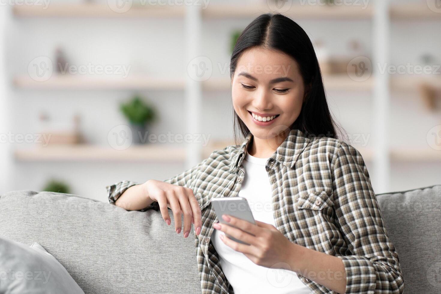 Cheerful asian lady sitting on sofa and using smartphone, chatting with girlfriend or lover, copy space. Pretty young woman with mobile phone chilling on couch, checking social media, home interior photo