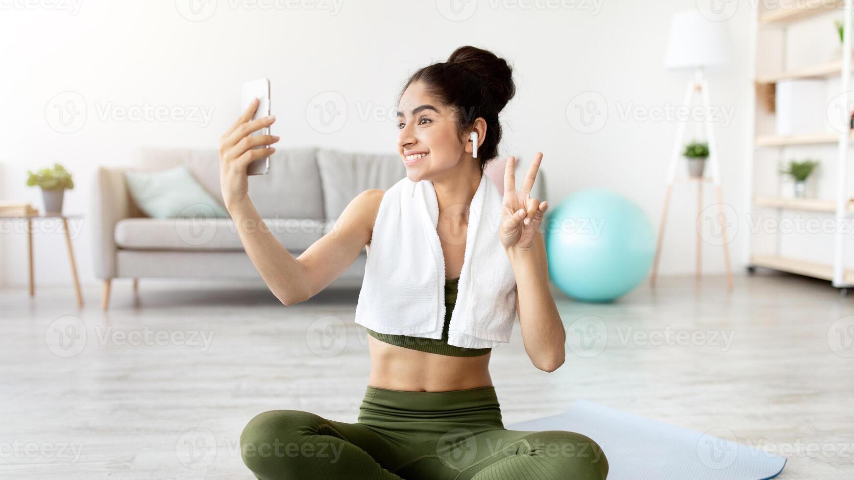 Joyful Indian woman taking selfie on break from home workout, full length. Happy Asian lady with smartphone making photo of herself on domestic training, making peace gesture and smiling at webcam