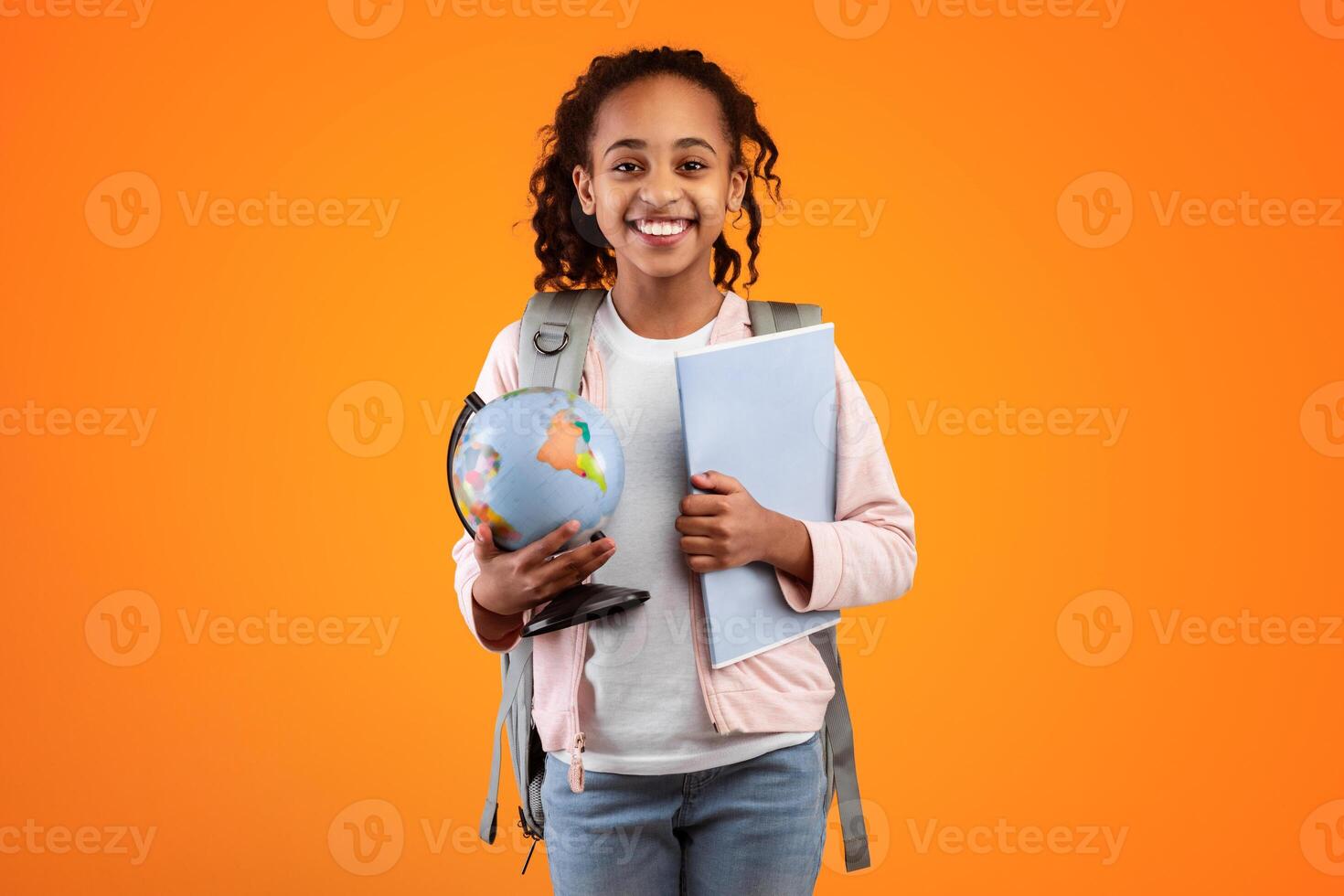 Portrait of cheerful young black girl holding Earth world globe and notebook, wearing backpack and looking at camera isolated on yellow color background at studio. Education in school concept photo
