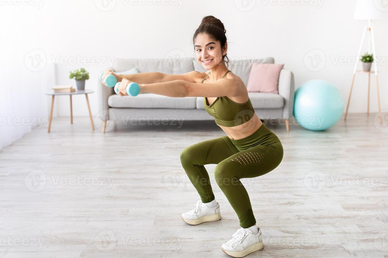 Strong young Indian woman working out with dumbbells, squatting at home, empty space. Fit Eastern lady taking care of her health, having domestic training, leading active lifesyle photo