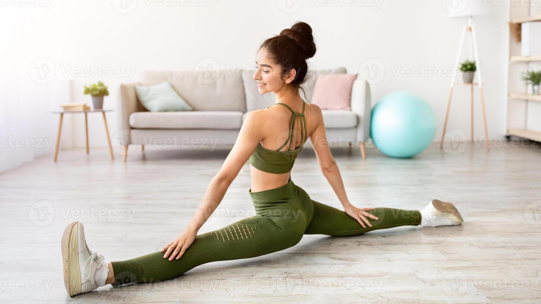 Sporty Indian woman sitting in splits, stretching legs at home, full length portrait. Charming Asian lady doing flexibility exercises, keeping active and strong during covid lockdown photo