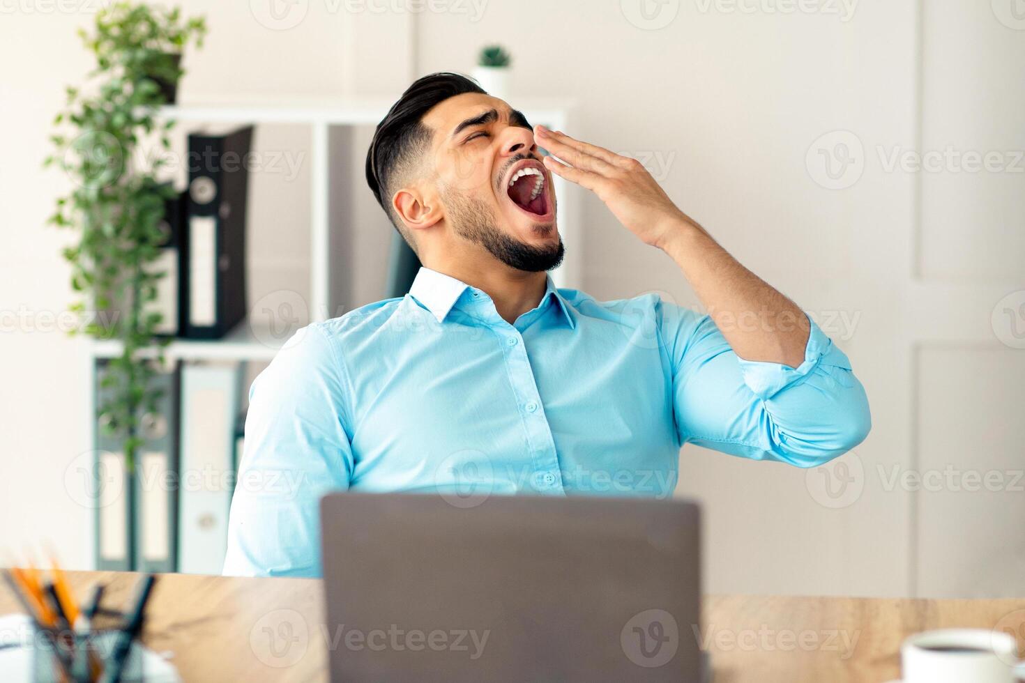 Sleepy young Arab guy yawning, tired of working with laptop at modern office. Millennial workaholic suffering from chronic fatigue, feeling sleepy or bored at workplace, copy space photo