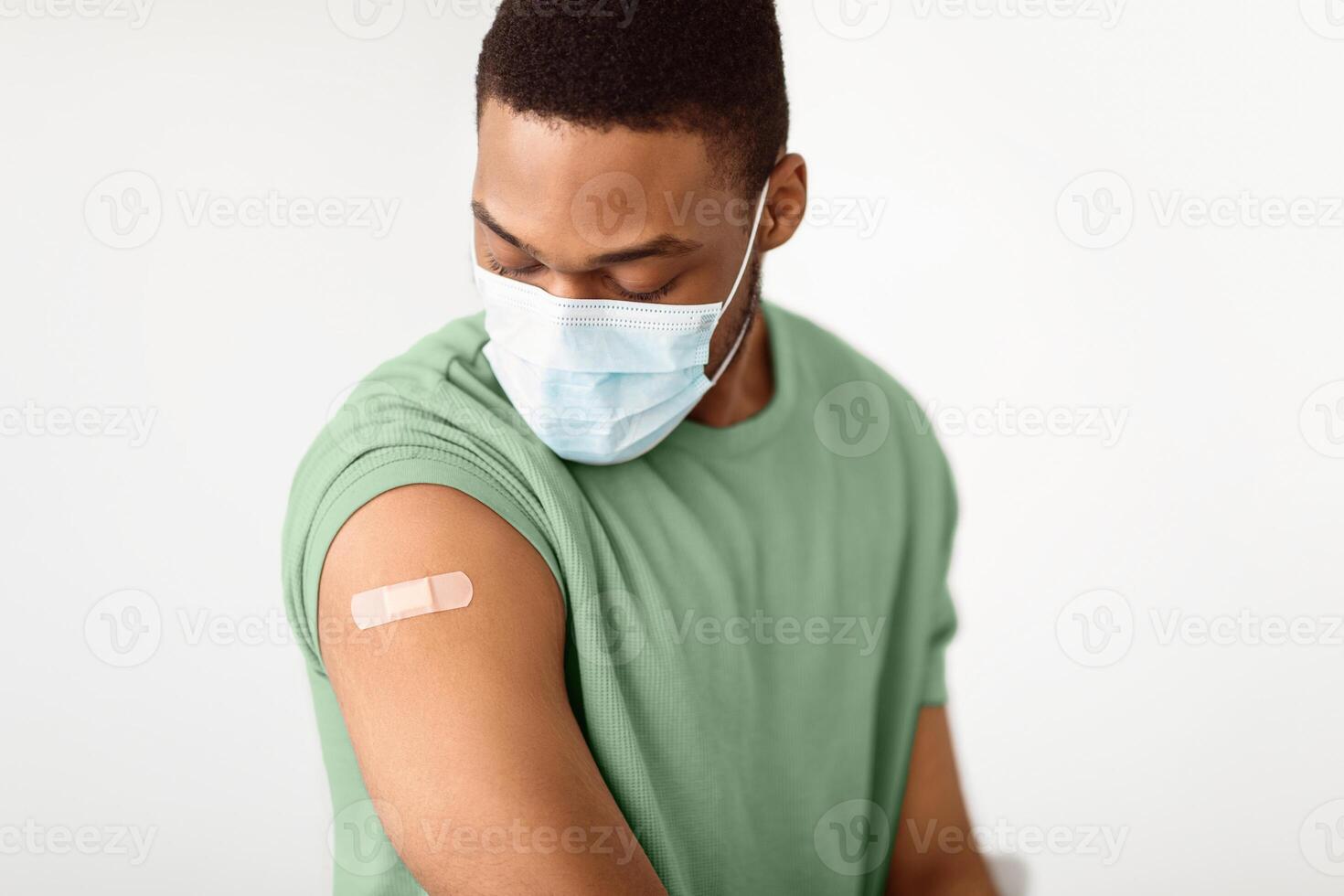 Covid-19 Coronavirus Vaccination. Vaccinated Black Man Looking At Arm With Injection Plaster Strip After Vaccine Shot Sitting On White Background, Wearing Protective Face Mask. Selective Focus photo