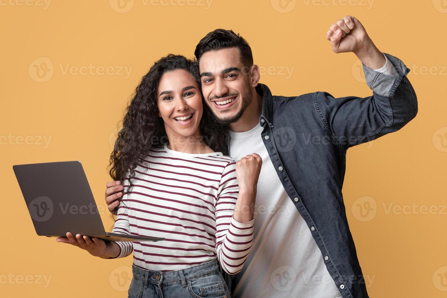 Online Win. Portrait Of Overjoyed Arabic Couple Celebrating Success With Laptop, Cheerful Middle Eastern Spouses Holding Computer, Raising Fists And Exclaiming With Excitement On Yellow Background photo