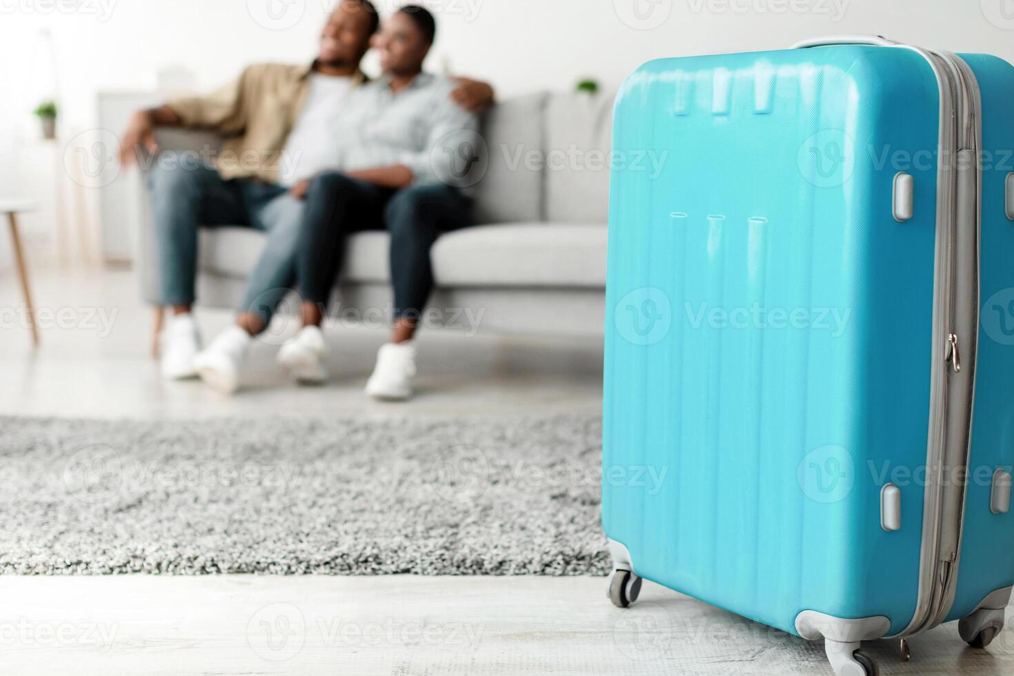 Blue Travel Suitcase Standing On Floor While Unrecognizable Black Couple Hugging Sitting On Couch Ready For Vacation Trip At Home. Tourists Waiting For Journey. Selective Focus On Luggage photo