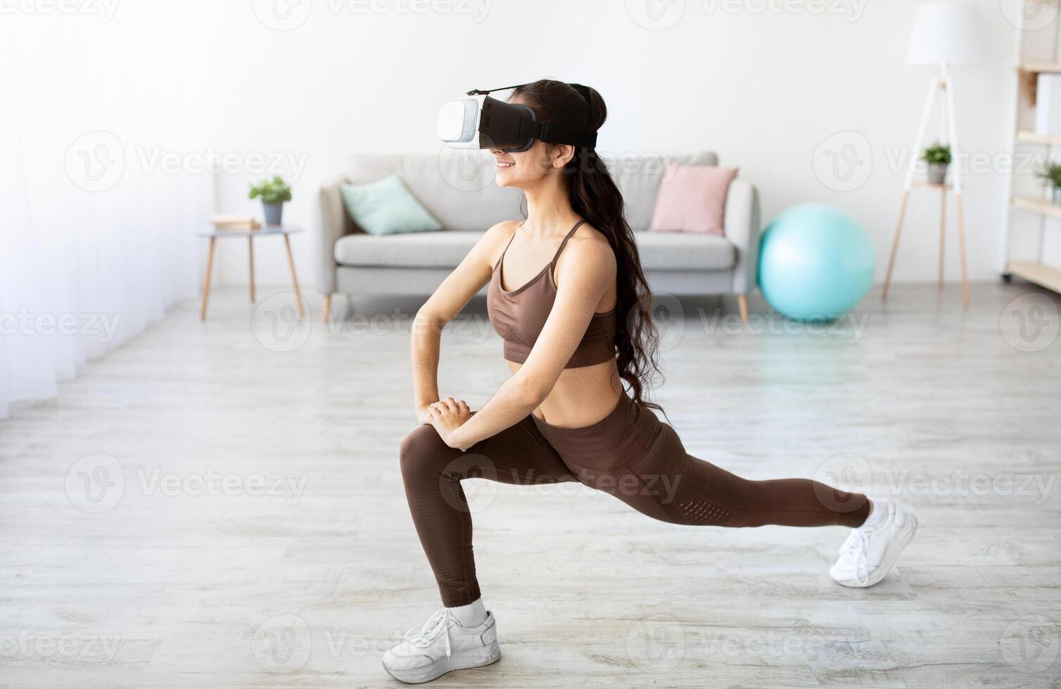 Athletic Indian lady wearing VR headset, doing virtual workout in augmented reality at home. Millennial Asian woman using contemporary technologies for online domestic training, copy space photo