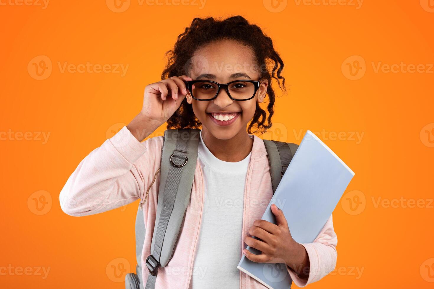 Closeup portrait of confident cheerful black female pupil wearing and touching eyeglasses, holding educational materials, posing and looking at camera isolated on yellow orange studio background wall photo
