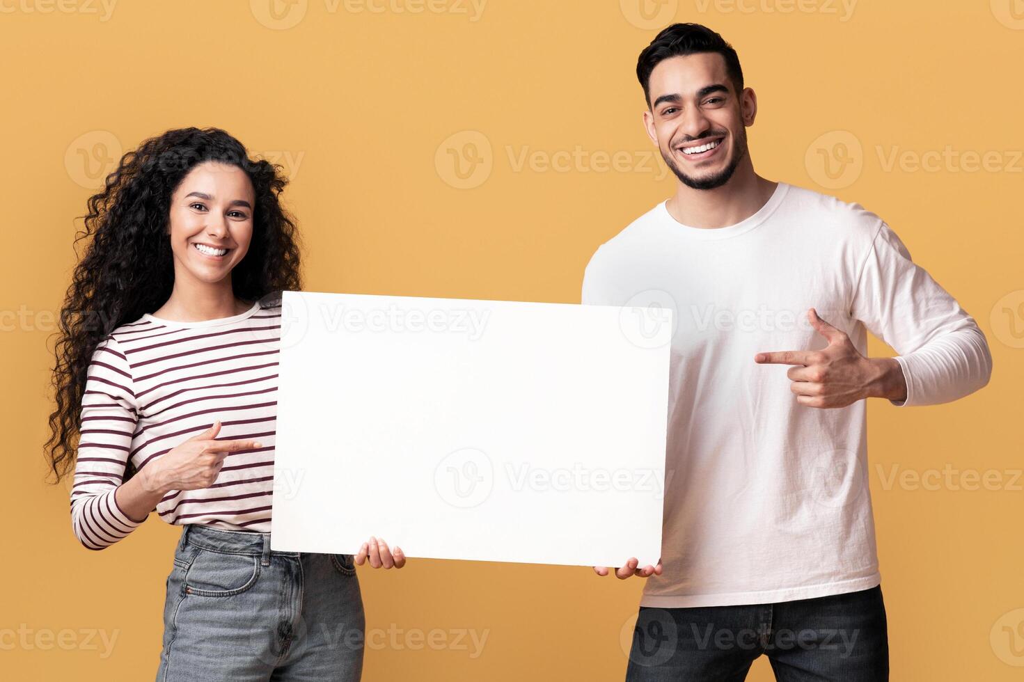 Place For Your Ad. Happy Middle Eastern Couple Pointing At White Blank Placard In Their Hands, Cheerful Young Arab Man And Woman Demonstrating Copy Space For Advertisement, Yellow Background photo
