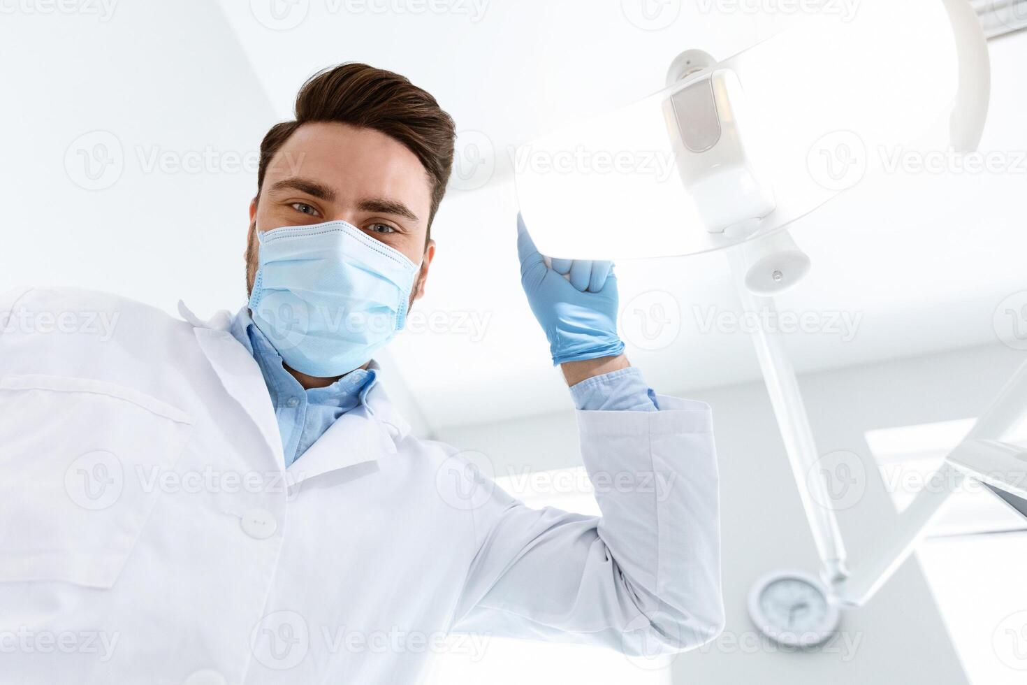 Male dentist in medical face mask turning on lamp, greeting patient in dental chair, looking at camera, copy space. Young man stomatologist beginning treatment, turning on light, closeup photo