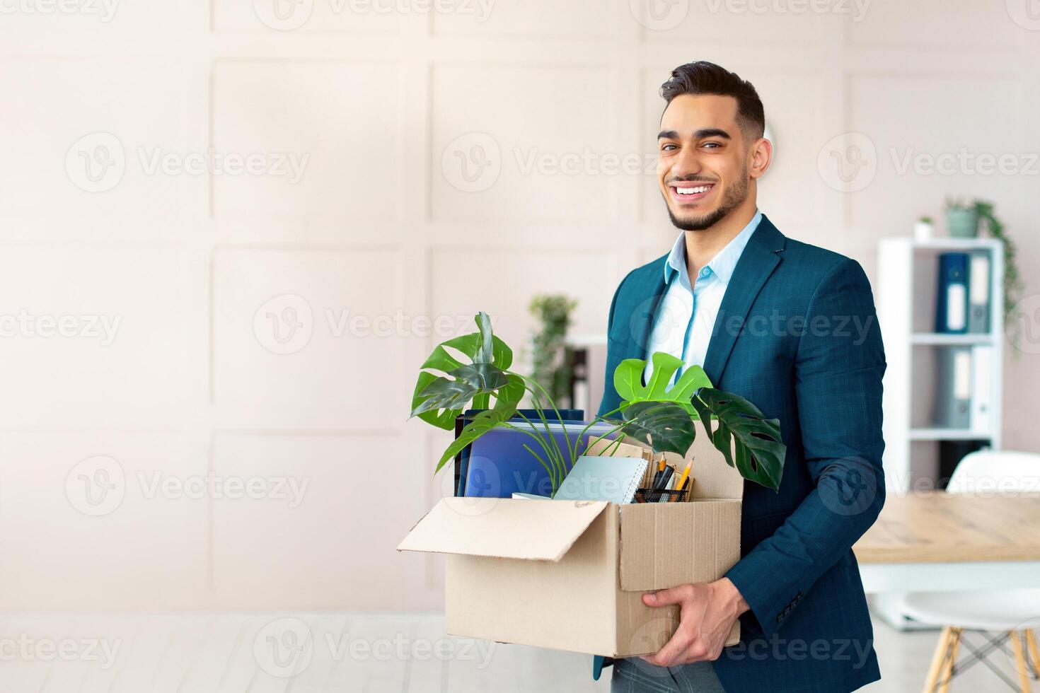 First day at office. Portrait of confident Arab businessman with belongings moving to new office. Happy young guy manager holding box with supplies, changing working place, indoors photo