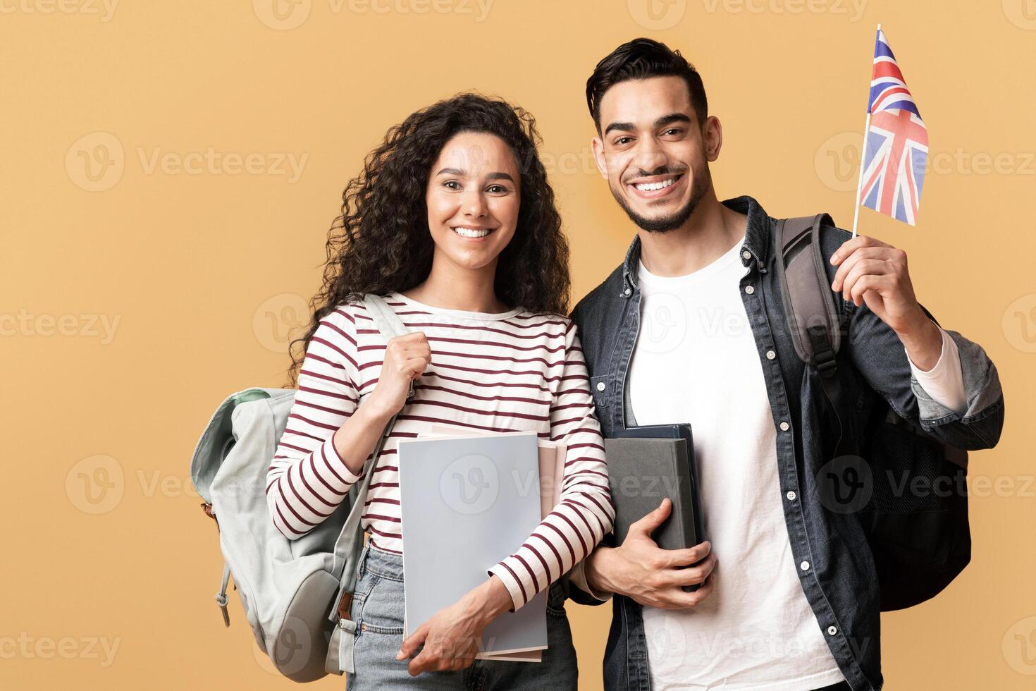 Study Abroad. Cheerful Young Arab Man And Woman With Backpacks Holding British Flag, Portrait Of Happy Smiling Students With Union Jack And Workbooks Over Yellow Studio Background, Copy Space photo