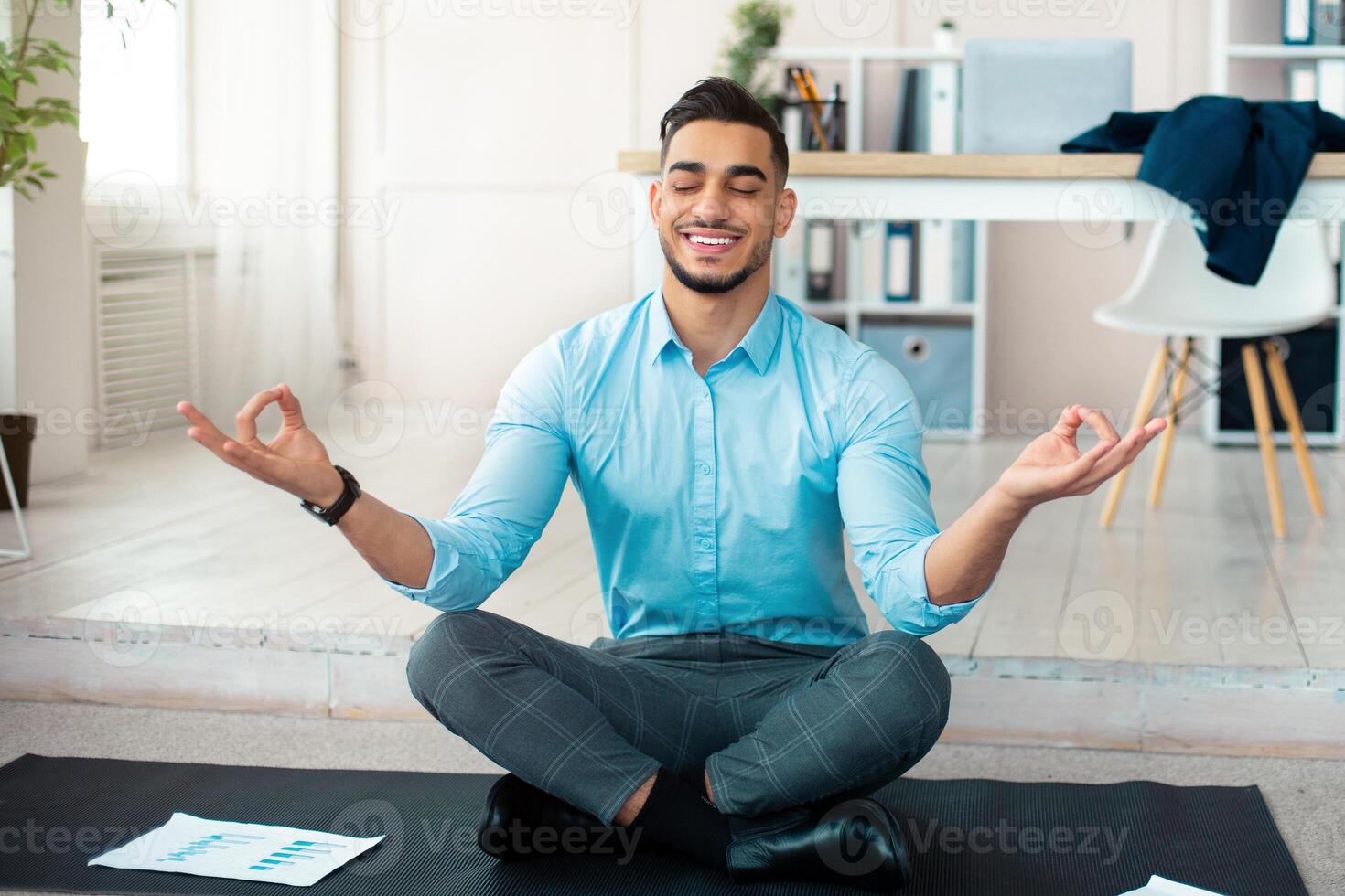 Workplace stress management concept. Calm Arab man meditating with closed eyes at modern office. Young businessman feeling peaceful and balanced, doing yoga on mat photo