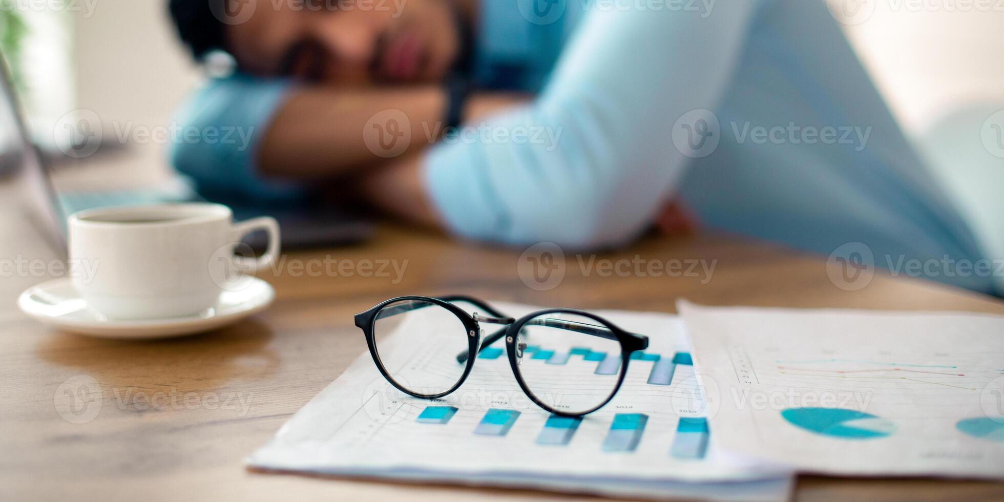 Exhausted Arab businessman sleeping on his office desk, tired of overworking, selective focus on documents and glasses. Millennial workaholic suffering from chronic fatigue at workplace, copy space photo
