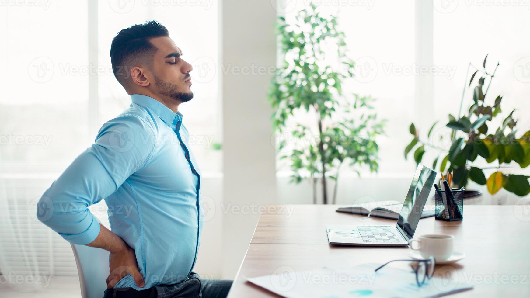 Young Arab businessman having acute back pain, massaging aching muscles, sitting at desk in modern office, copy space. Millennial guy having health problem, bad posture due to sedentary work photo