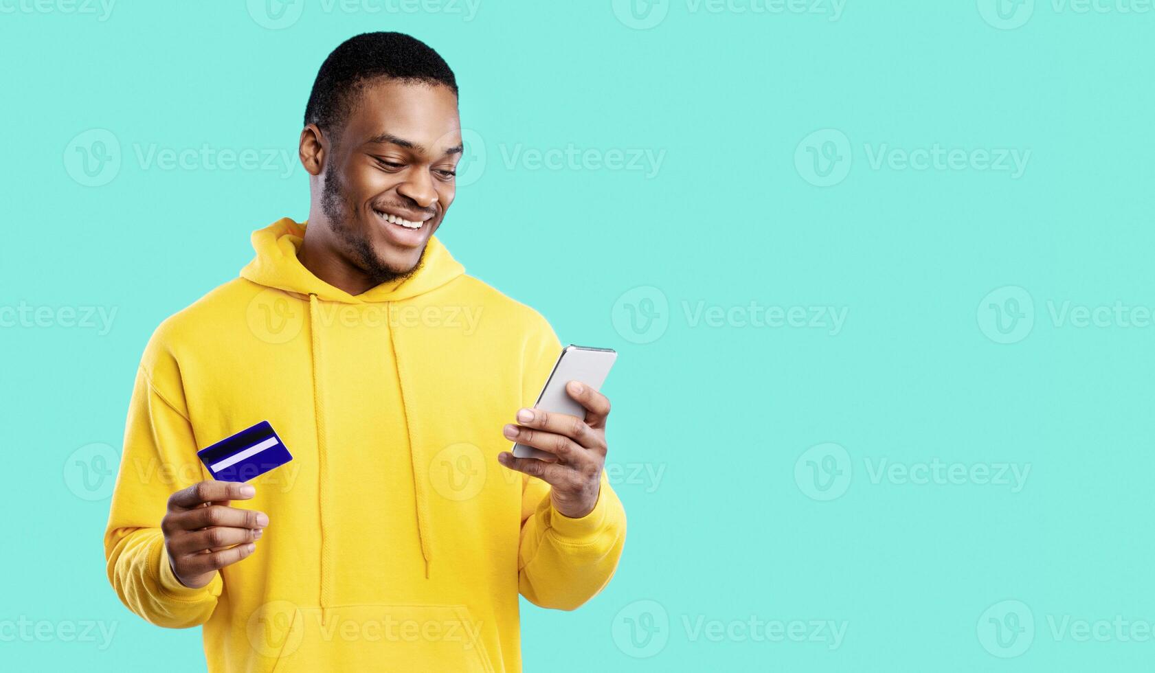 Mobile Shopping. Happy African American Man Using Credit Card And Cellphone Purchasing Something In Online Shop Application Standing Over Blue Studio Background. E-Commerce Concept photo