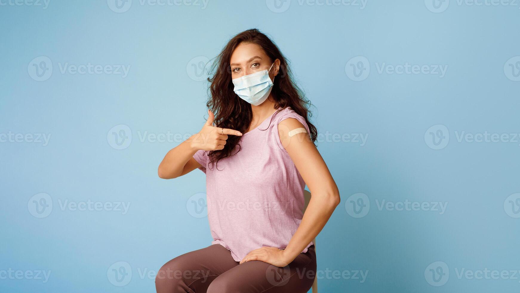 Antiviral Vaccination. Vaccinated Woman Wearing Face Mask Pointing At Arm Showing Plaster After Covid-19 Vaccine Injection Sitting On Blue Background. Coronavirus Immunization. Panorama, Free Space photo