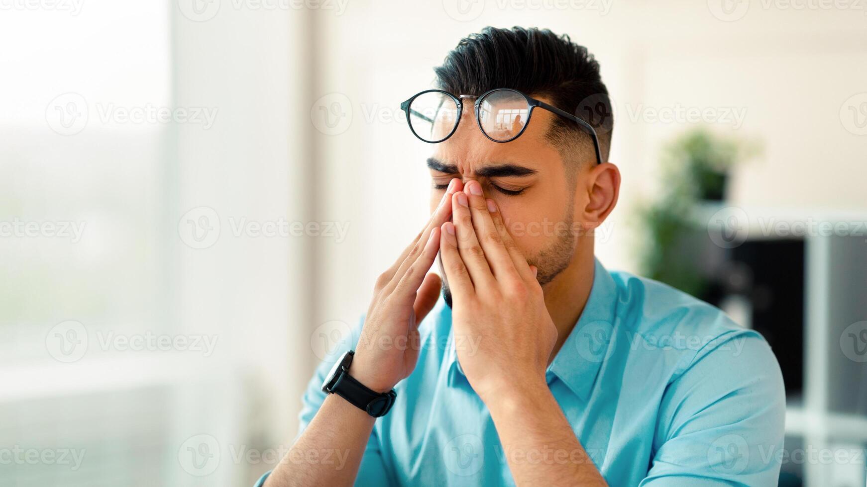 Young Arab office worker in glasses rubbing tired eyes, exhausted from overworking at modern office, copy space. Young Eastern businessman feeling eye fatigue or pain indoors photo