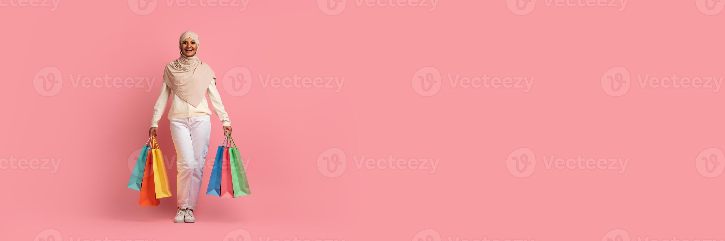 Cheerful young muslim woman walking with shopping bags over pink studio background, panorama with copy space photo