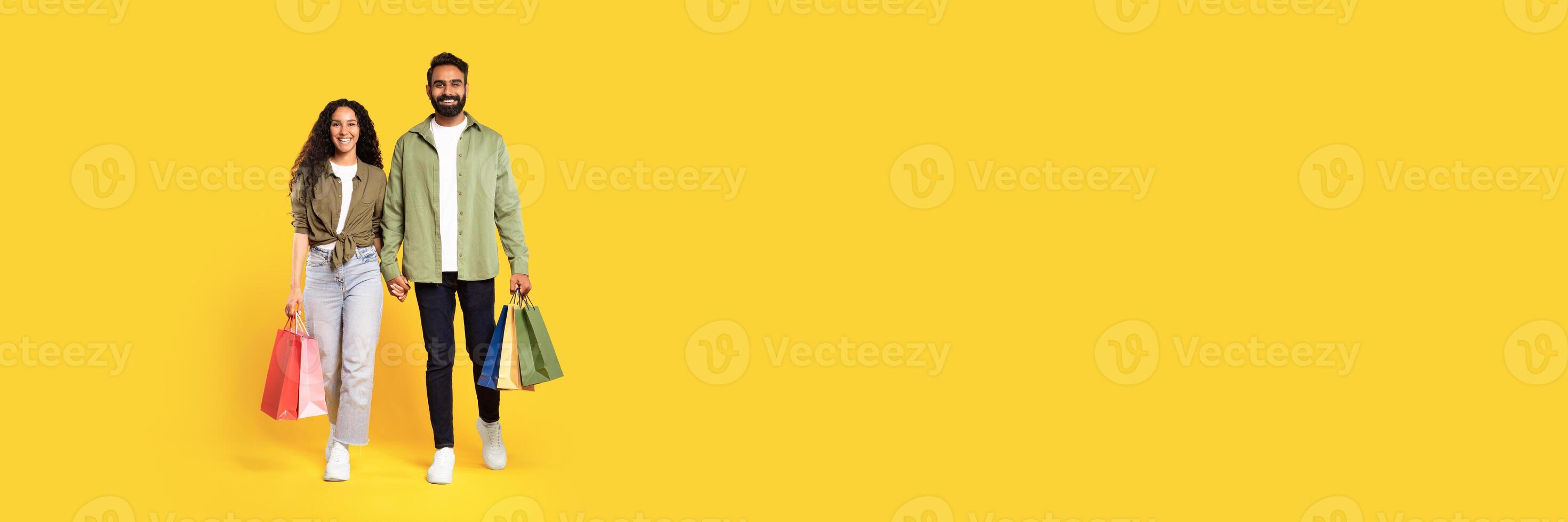 Shopping and consumerism. Happy arabic young man and woman in casual holding colorful shopping bags, walking holding hands and smiling at camera over yellow background. Sales season, retail offer photo