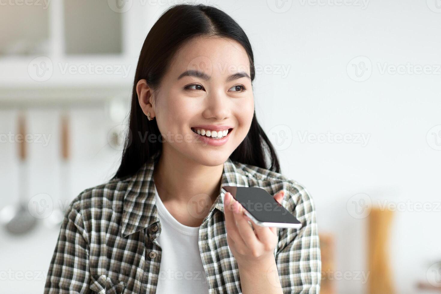 Smiling asian woman recording voice message on mobile phone while eating oatmeal at cozy white kitchen, looking at copy space, having digital tablet on table. Mobile application for communication photo