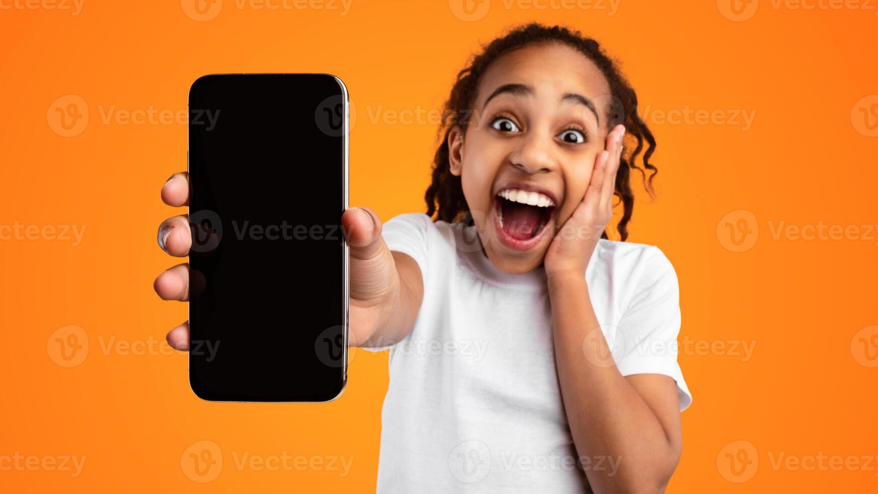 Wow, Amazing Offer. Portrait of excited African American girl holding mobile phone with empty screen in hand, showing device close to camera and touching cheek, mock up, yellow studio background photo