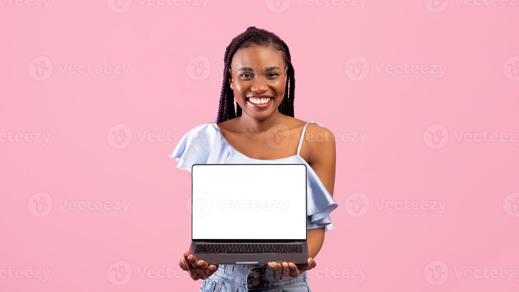 Funky black lady holding laptop pc with blank screen for your ad or website on pink studio background. Cool Afro woman showing portable computer with mockup for design or advertisement photo