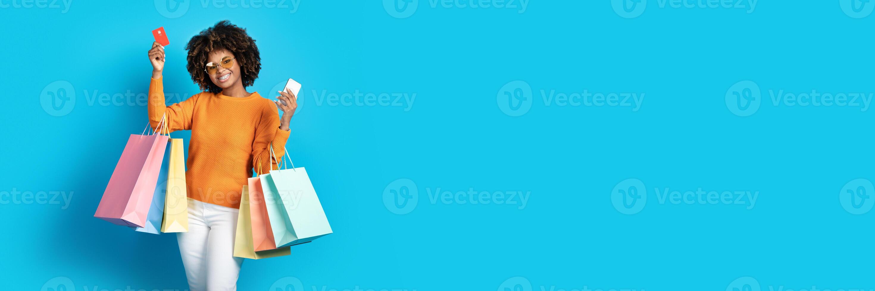 Pretty young African American woman with colorful shopping bags holding credit card and smartphone over blue studio background, panorama with copy space. E-commerce concept photo