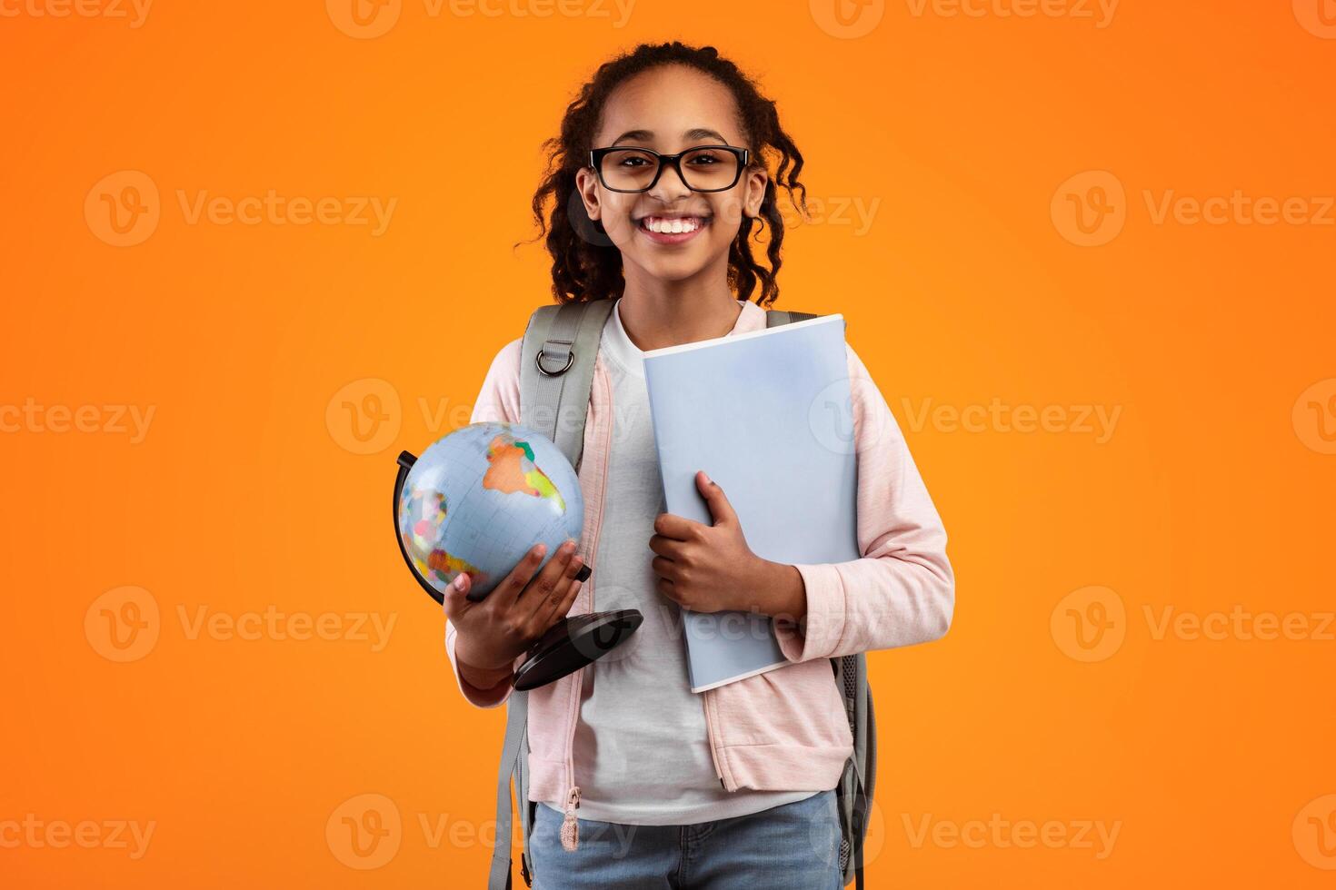 Portrait of cheerful African American girl in glasses holding Earth world globe and notebook, wearing backpack and looking at camera isolated on yellow studio background. Education in school concept photo