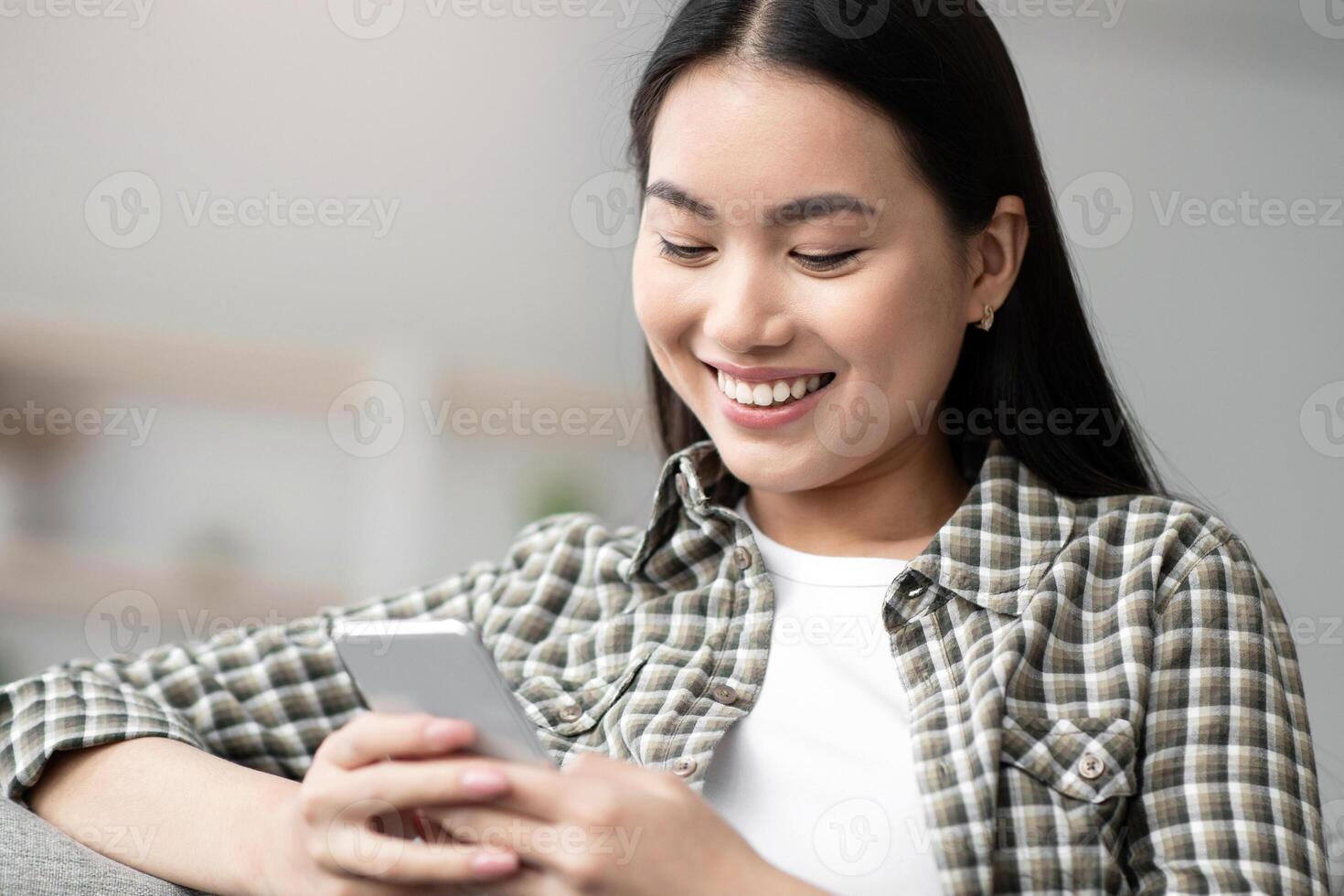 Cheerful asian lady sitting on sofa and using smartphone, chatting with girlfriend or lover, copy space. Pretty young woman with mobile phone chilling on couch, checking social media, closeup photo