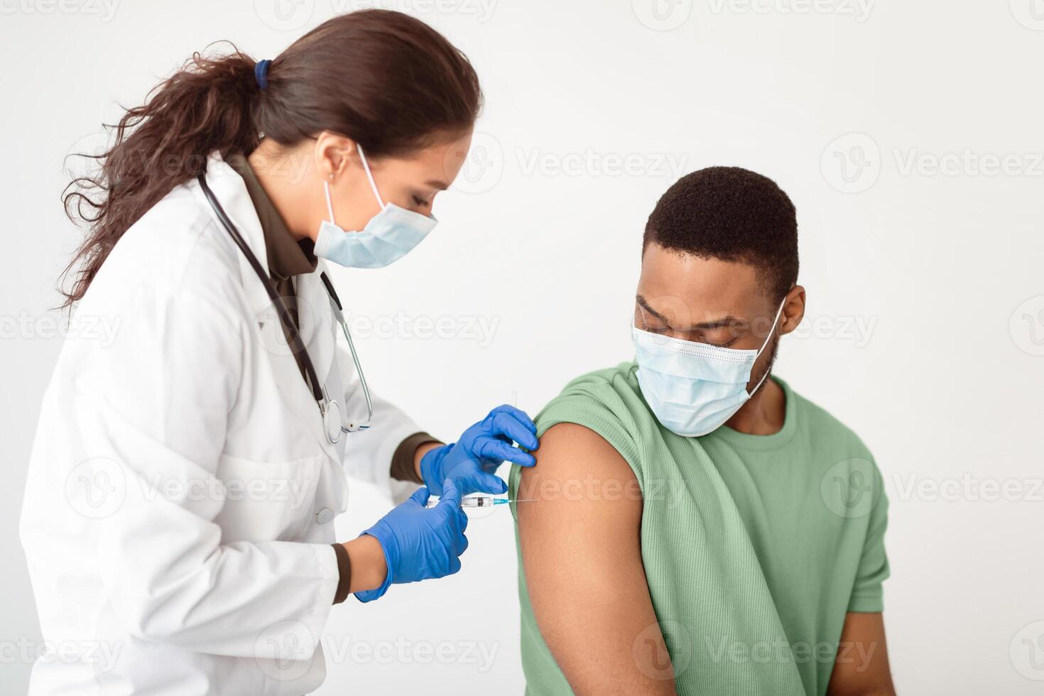 Covid-19 Vaccination. General Practitioner Vaccinating African American Male Patient Over White Studio Background, Applying Adhesive Plaster On Arm After Giving Vaccine Injection To Guy In Face Mask photo