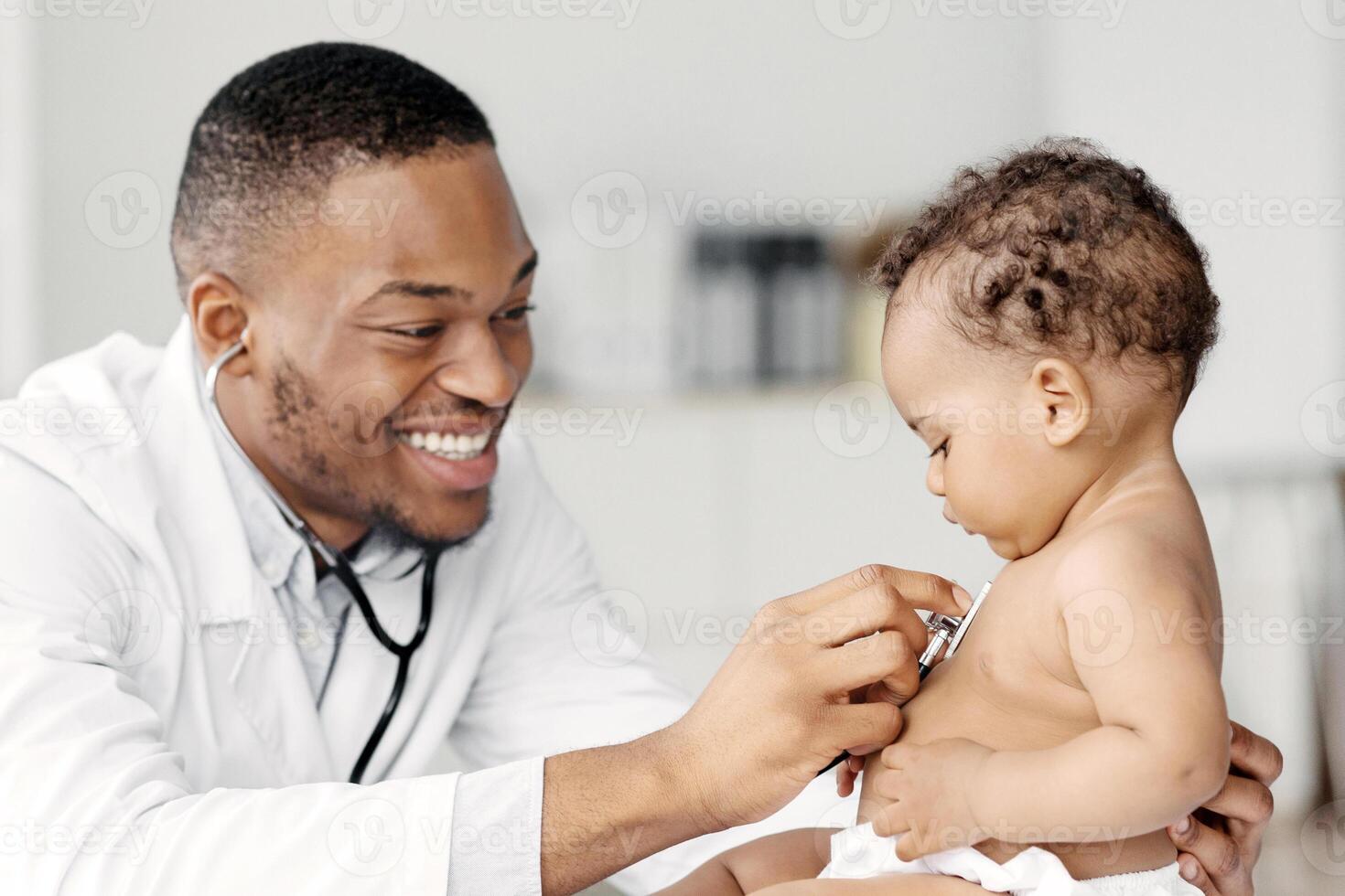 Black Pediatrician Doctor Doing Check Up To Cute Newborn Baby In Diaper, African American Pediatrist Using Stethoscope For Listening Child's Heartbeat, Checking Infant's Heart And Lungs In Clinic photo